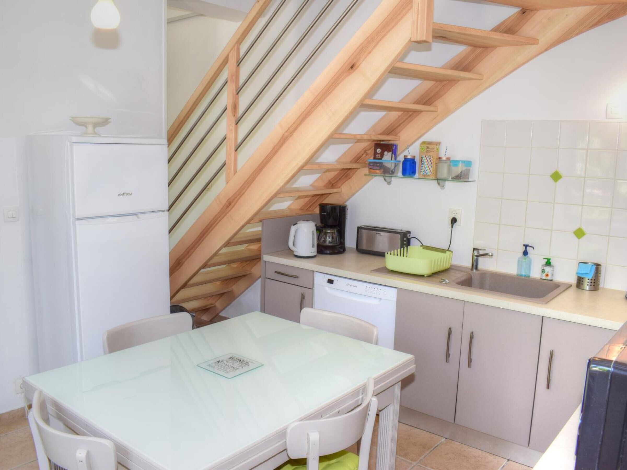 A compact kitchen with a dining area, wooden staircase, and a white refrigerator.