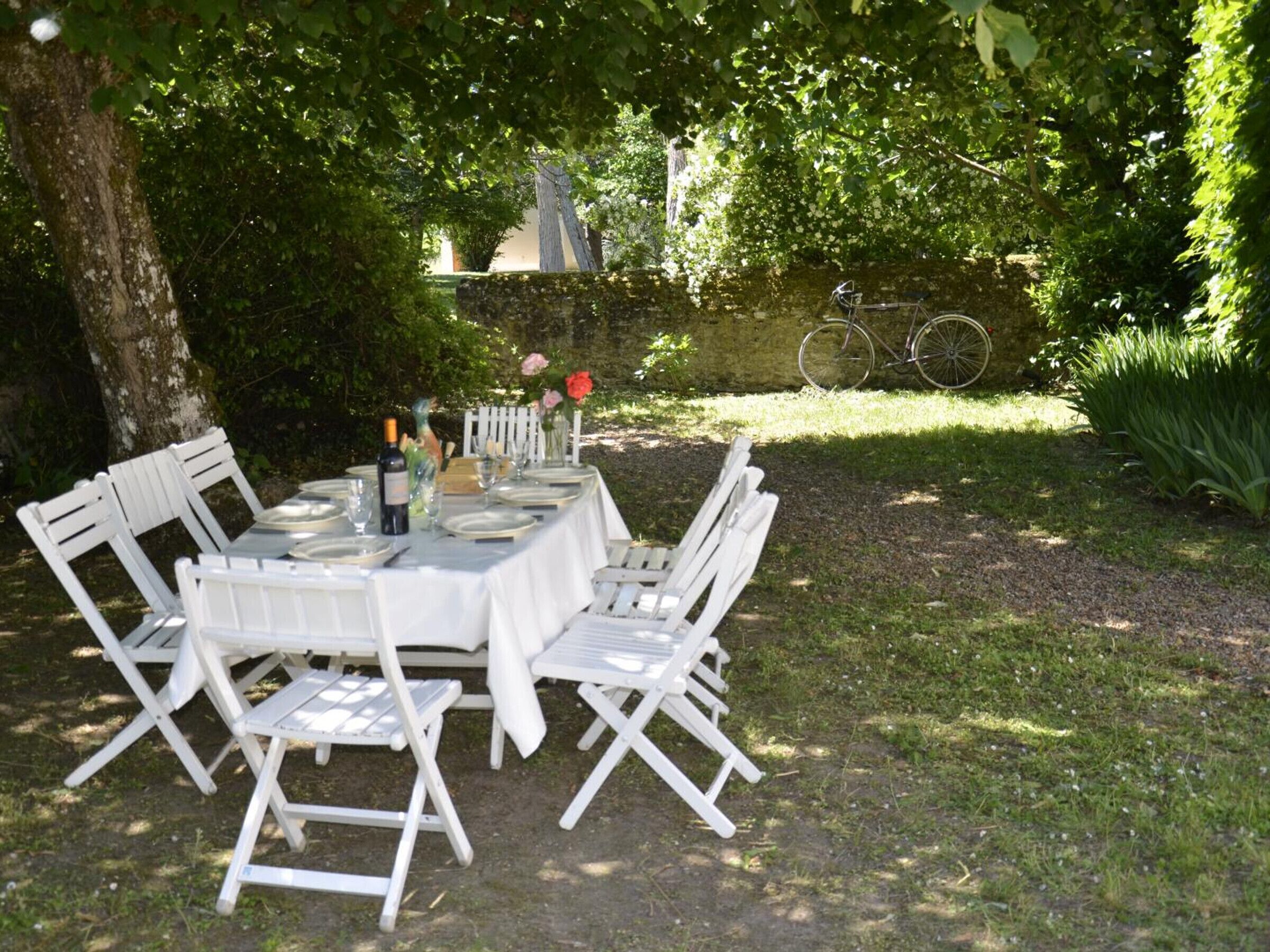 Ein Essbereich im Freien mit einem Tisch für vier Personen, umgeben von weißen Stühlen, in einem Garten mit Bäumen und einer Steinmauer im Hintergrund.