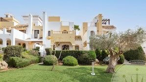 A multi-story building with a Mediterranean architectural style, featuring balconies, a well-maintained garden with various shrubs and a large tree, and a stone pathway leading to the entrance. - La Manga Club Monte Claro Townhouse 606 (Atamaría)