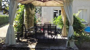 A covered patio with a table and chairs, surrounded by greenery and a white building in the background. - One-story house with park near Beauval Zoo (Meusnes)