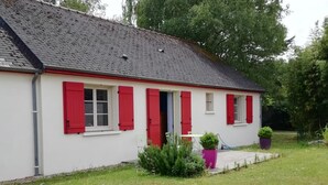 A white building with red shutters, a grey roof, and a small garden area with potted plants.