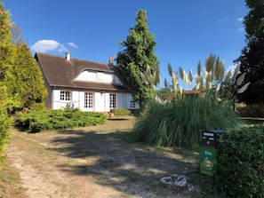Une maison blanche avec un toit foncé, entourée de verdure et de plantes de la famille des graminées.