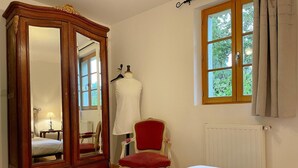Un dormitorio con un armario grande de madera, una butaca tapizada de rojo, una cama con colcha estampada y una ventana con vistas a zonas verdes.