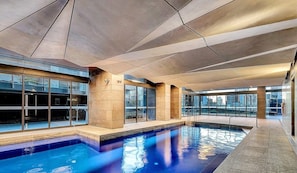 Piscina interior moderna com paredes de vidro transparente e design geométrico no teto.