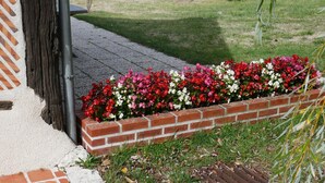Un jardinier en briques orné de fleurs colorées, un chemin en béton et une zone herbeuse avec des arbustes.