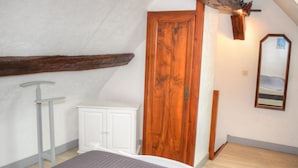 Une chambre avec un plafond en pente, une porte en bois, une armoire blanche, un miroir et un lit avec un matelas gris.
