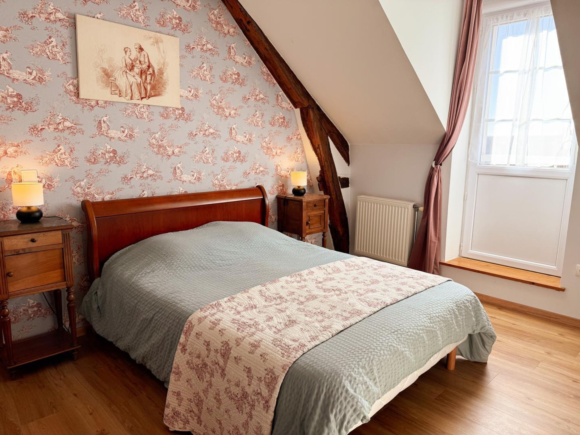 Une chambre à coucher avec un lit en bois, une table de chevet, une lampe, un radiateur et une fenêtre avec des rideaux.