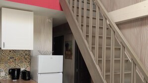Una cocina con un refrigerador blanco, una cafetera y un lavavajillas. Hay una escalera con barandilla de madera que lleva a otra habitación.