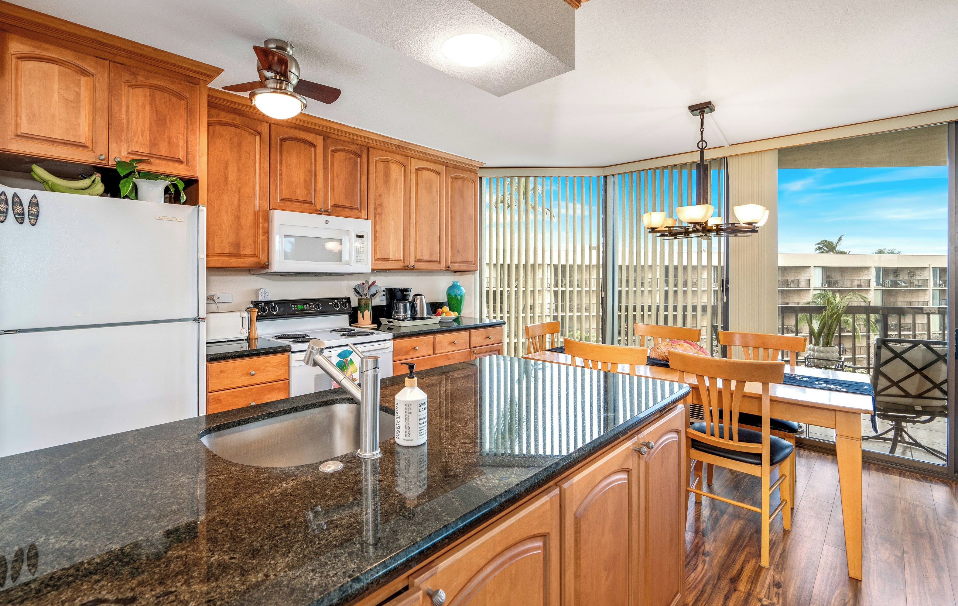 A kitchen with wooden cabinets, a granite countertop, a refrigerator, a microwave, and a dining area with a table and chairs.