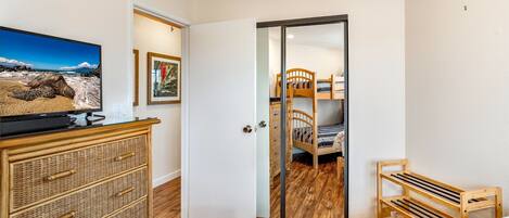 A bedroom with a bed, a wooden dresser, a television, and a door leading to a bathroom with a wooden bench.