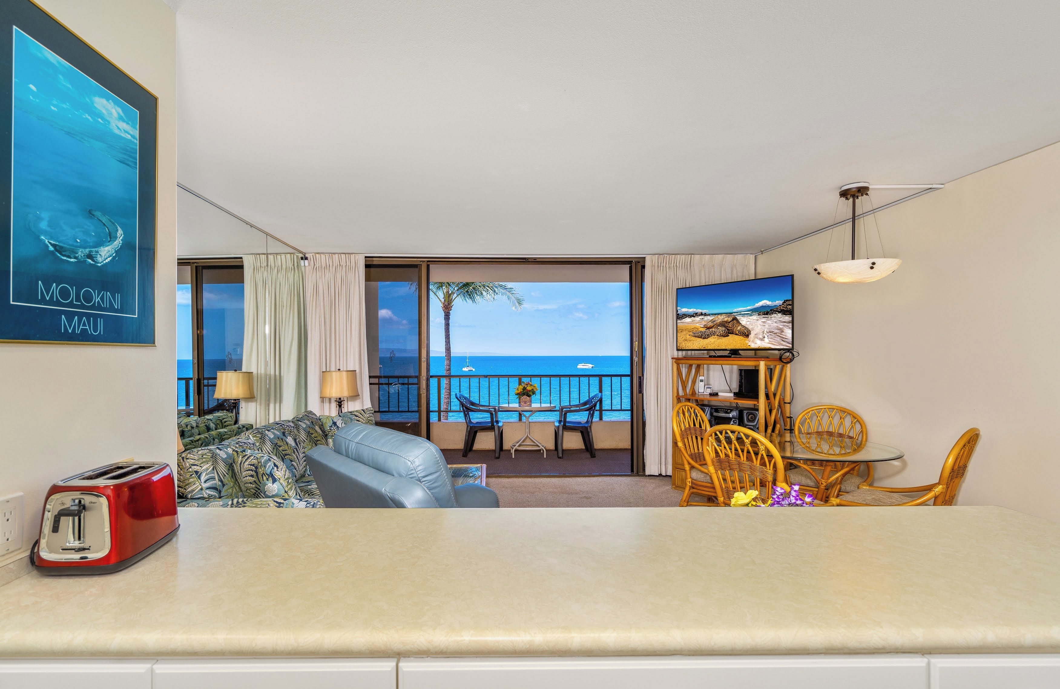 Habitación de hotel con balcón, sofá, mesa de comedor con sillas y un cuadro de una playa.