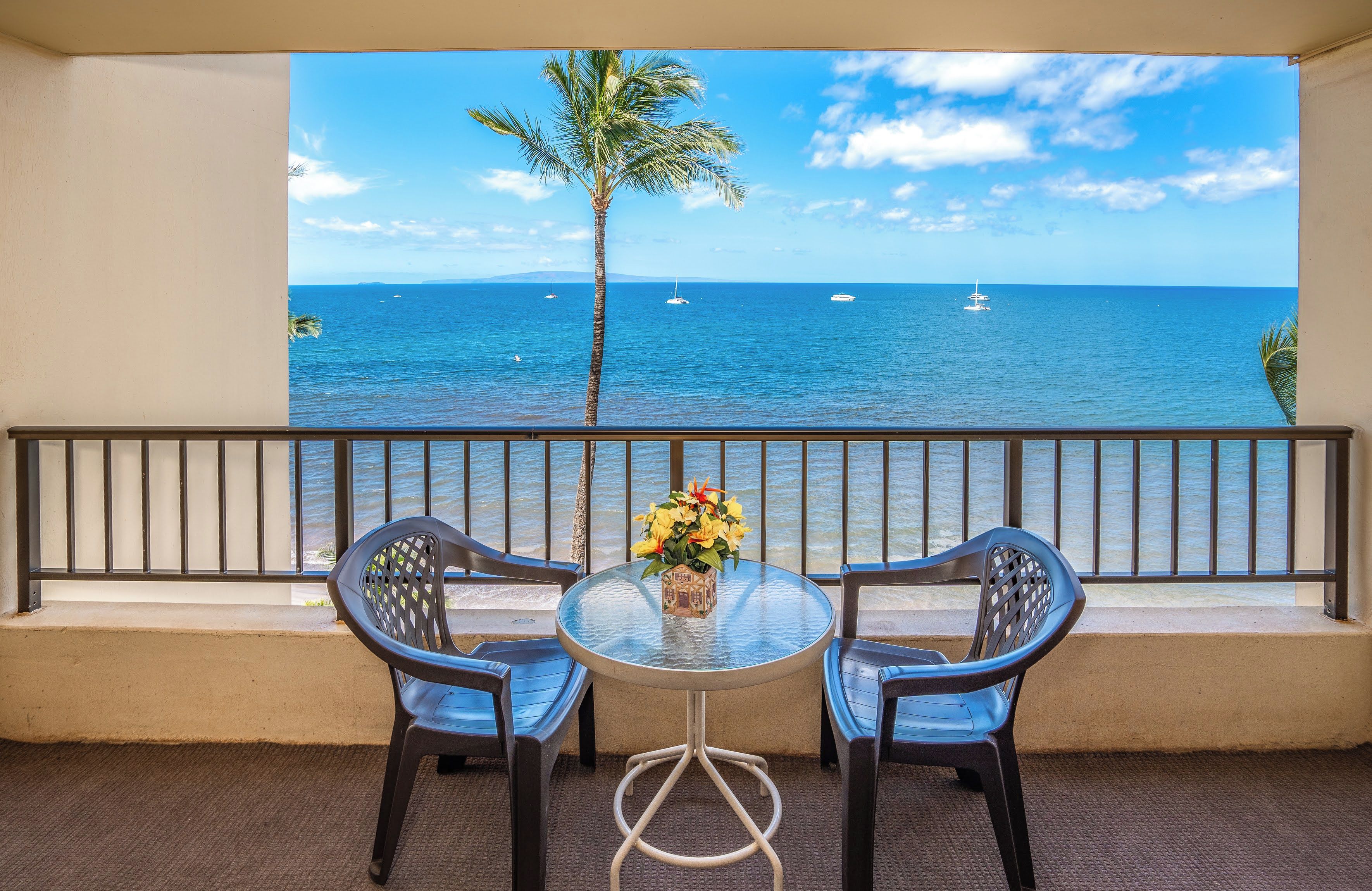 Varanda com duas cadeiras e uma mesa, com vista para o mar azul e límpido, onde se veem barcos e uma palmeira.