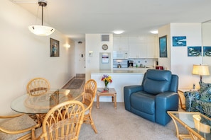 Een moderne woonkamer met een glazen salontafel, rieten stoelen, een blauwe leren bank en een keuken op de achtergrond.
