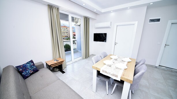 A modern living room with a sofa, a dining table set for four, and a sliding glass door leading to an outdoor area. - Alkan Apart (Demre)