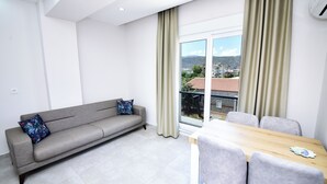 A modern living room with a sofa, a dining table with chairs, and a large window with curtains. - Alkan Apart (Demre)