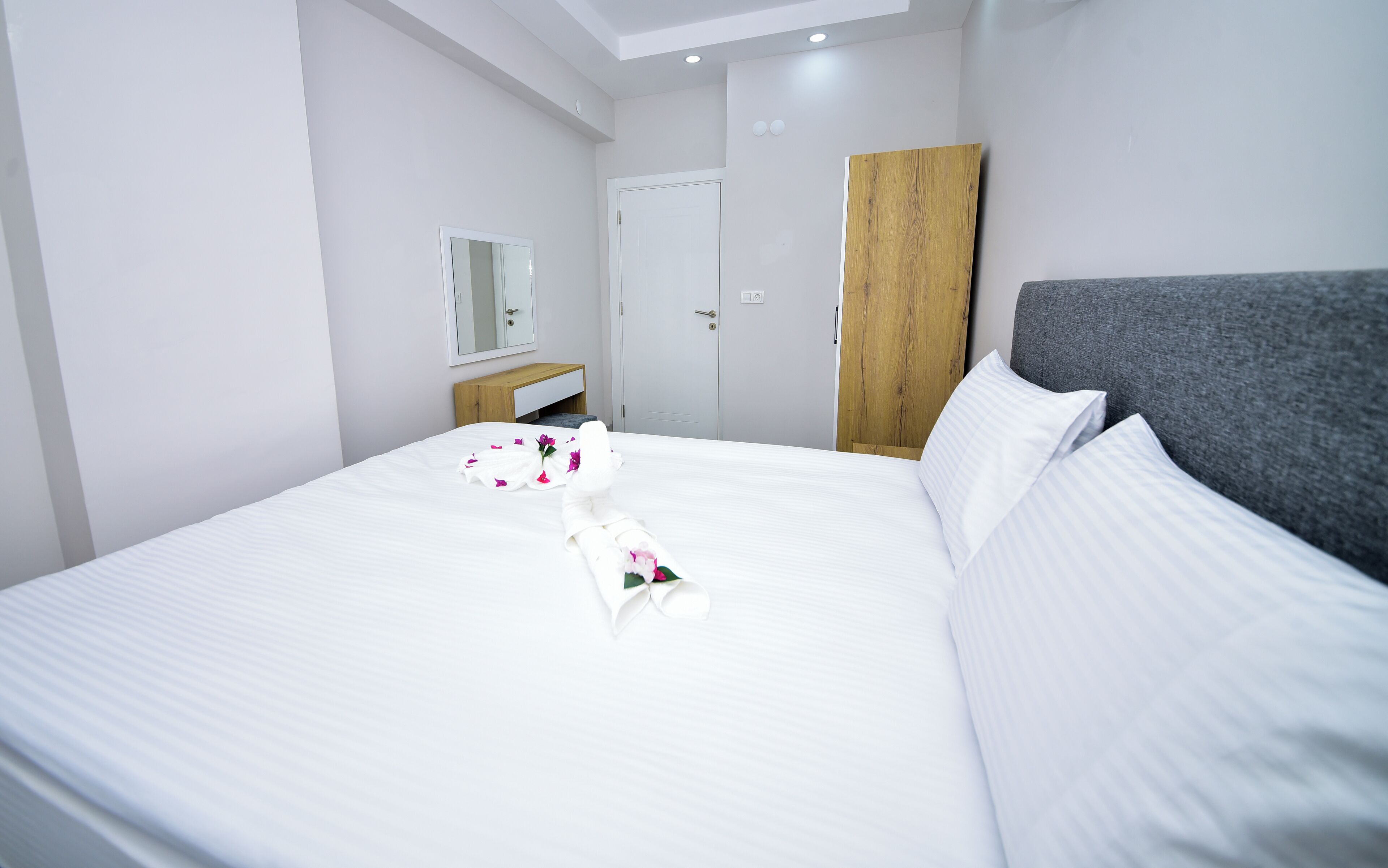 A neatly made bed with white linens and a floral arrangement, a wooden nightstand, a grey upholstered headboard, and a white wardrobe.