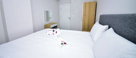 A neatly made bed with white linens and a floral arrangement, a wooden nightstand, a grey upholstered headboard, and a white wardrobe.