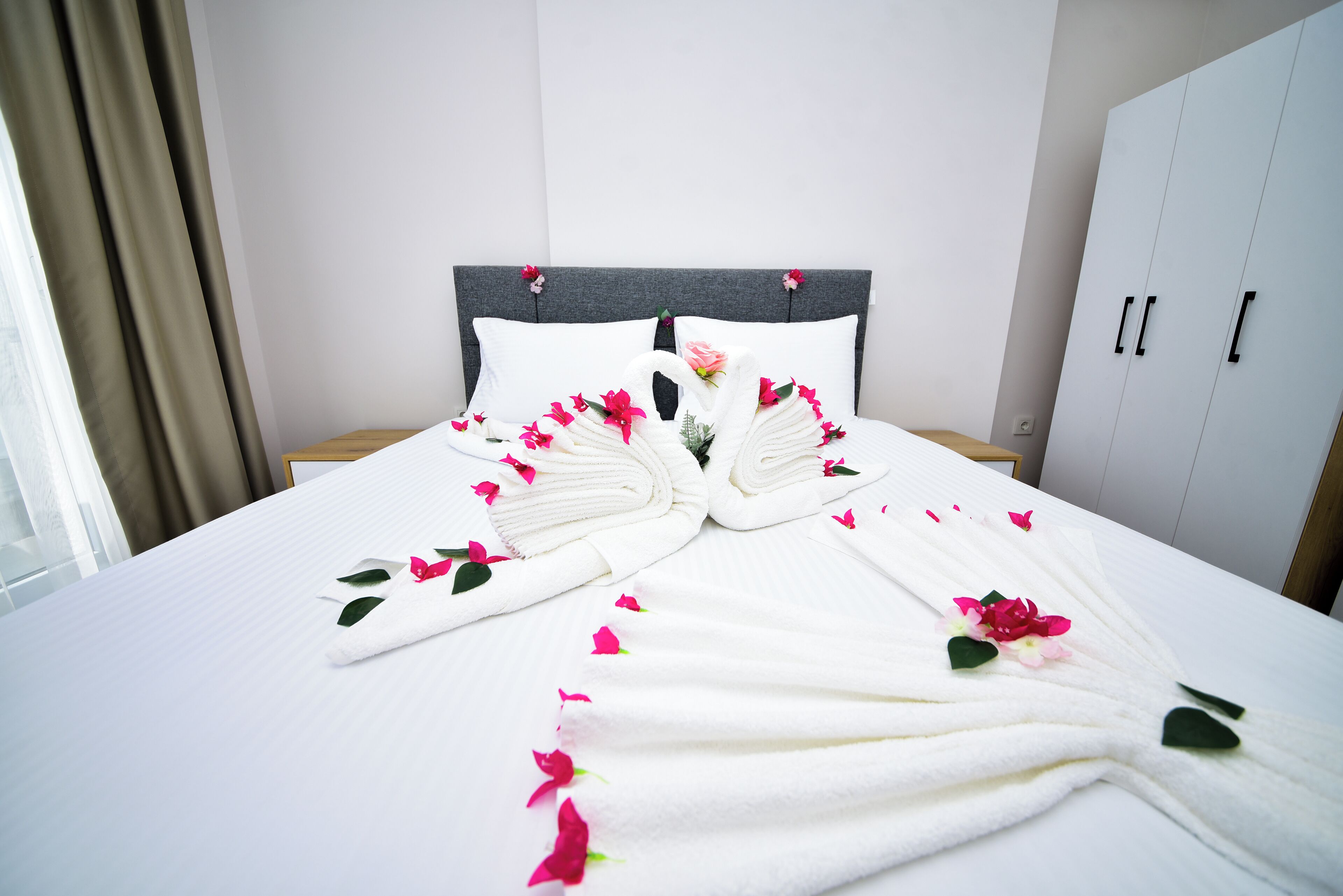 A neatly made bed with white linens, pink flower petals, and a swan-shaped towel arrangement.