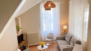 Un salon moderne avec un canapé gris, des chaises blanches, une petite table ornée de fleurs, un radiateur et un luminaire suspendu.