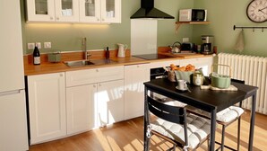 Une cuisine compacte avec des meubles blancs, un plan de travail en bois, une hotte noire, un four à micro-ondes, une bouilloire et une table à manger avec des chaises.