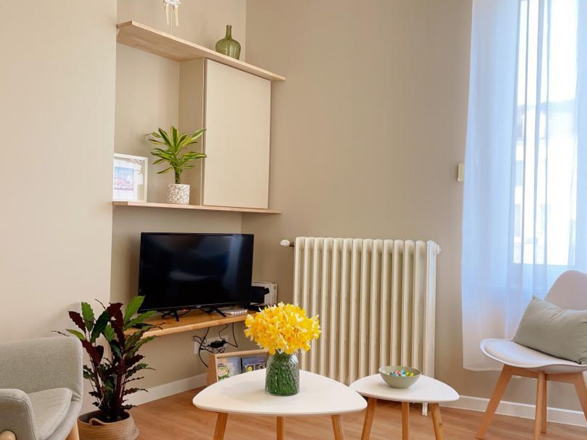 A modern living room with a flat-screen TV, a small white table with a vase of yellow flowers, a chair, and a shelf with decorative items.