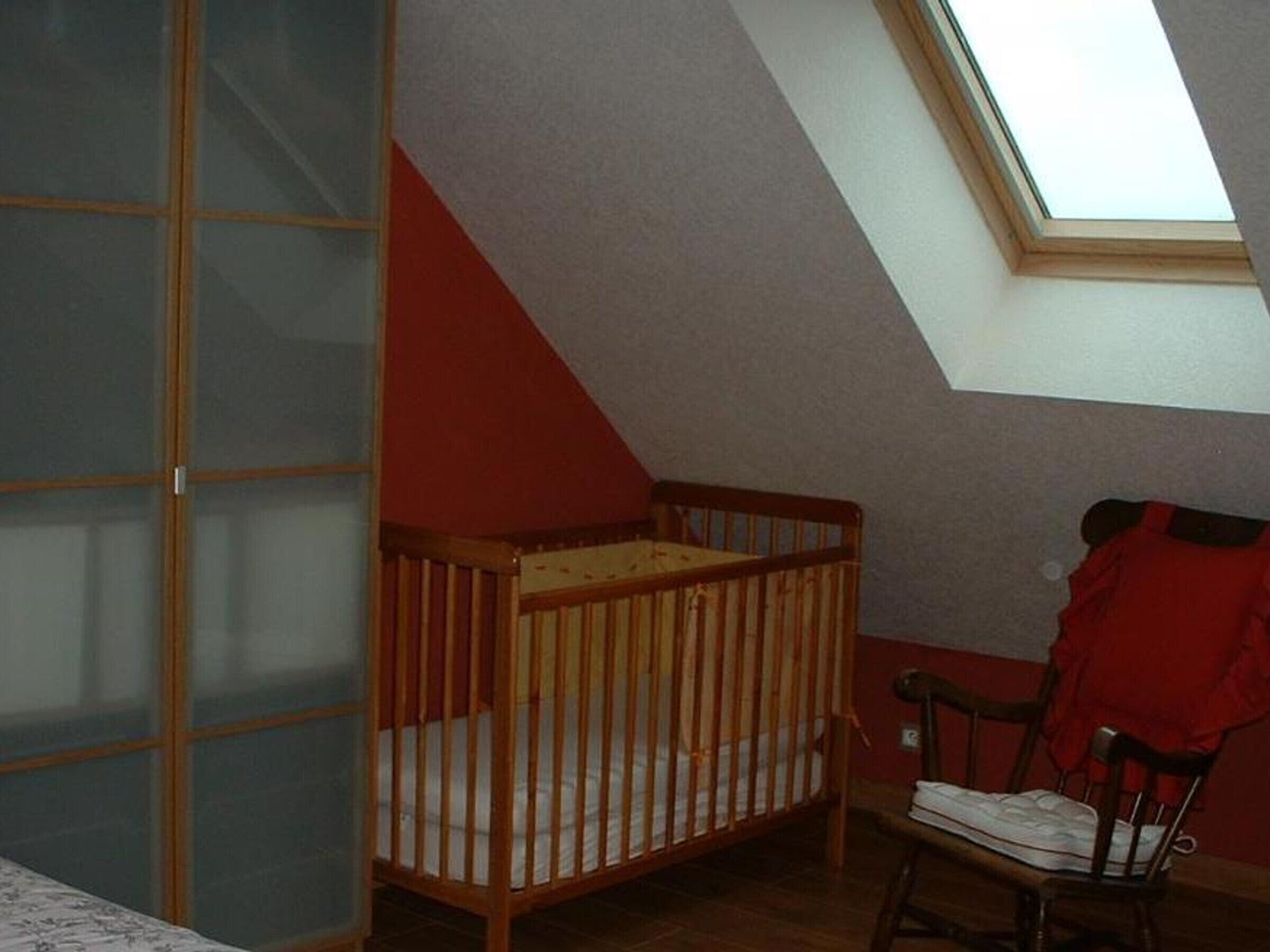 A room with a sloped ceiling, a wooden crib, a chair, and a window.