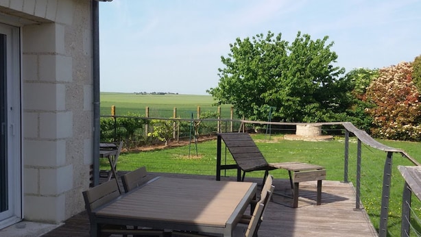 Une terrasse en bois avec une table et des chaises, un mur en pierre et une vue sur un champ verdoyant et des arbres.