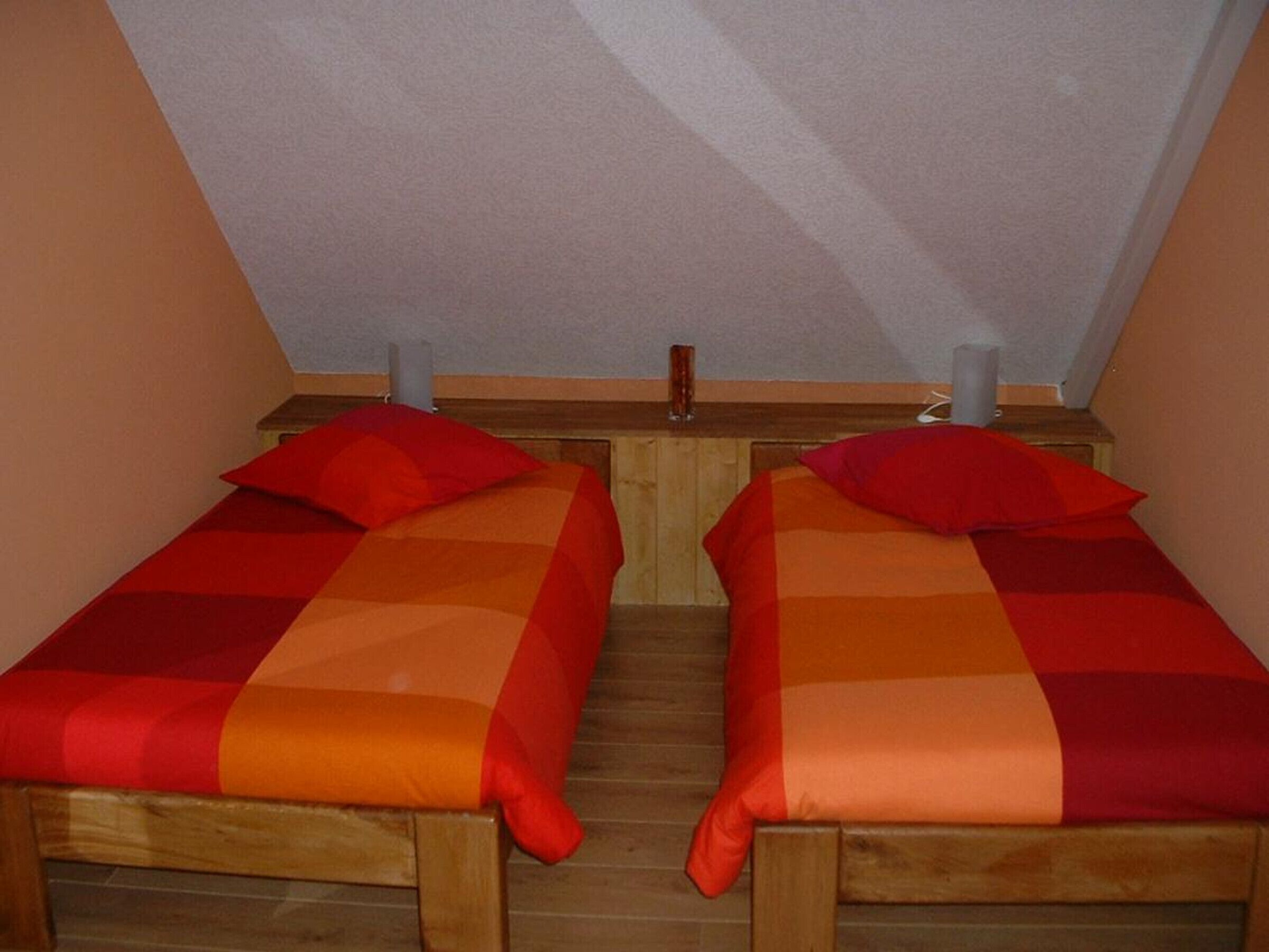 Twee houten bedden met rode en oranje beddengoed in een kamer met schuine plafonds.