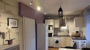 A compact kitchen with a refrigerator, microwave, oven, and washing machine. There is a dining table with a pink placemat and a potted orchid.