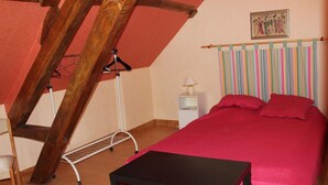 A bedroom with a sloped ceiling, a bed with a red bedspread, a small bedside table with a lamp, and a wooden chair.
