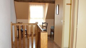 A narrow hallway with a wooden staircase leading to an upper level. There is a window with curtains, a chair, and a table in the distance.