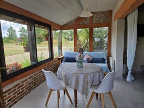 A room with a brick wall, a wooden ceiling, a round table with chairs, a sofa, and large windows with a view of greenery. - Charming Restored Barn with Tennis, Near Loire Castles & ZooParc de Beauval, Pet-Friendly, Sleeps 3 (Cheverny)