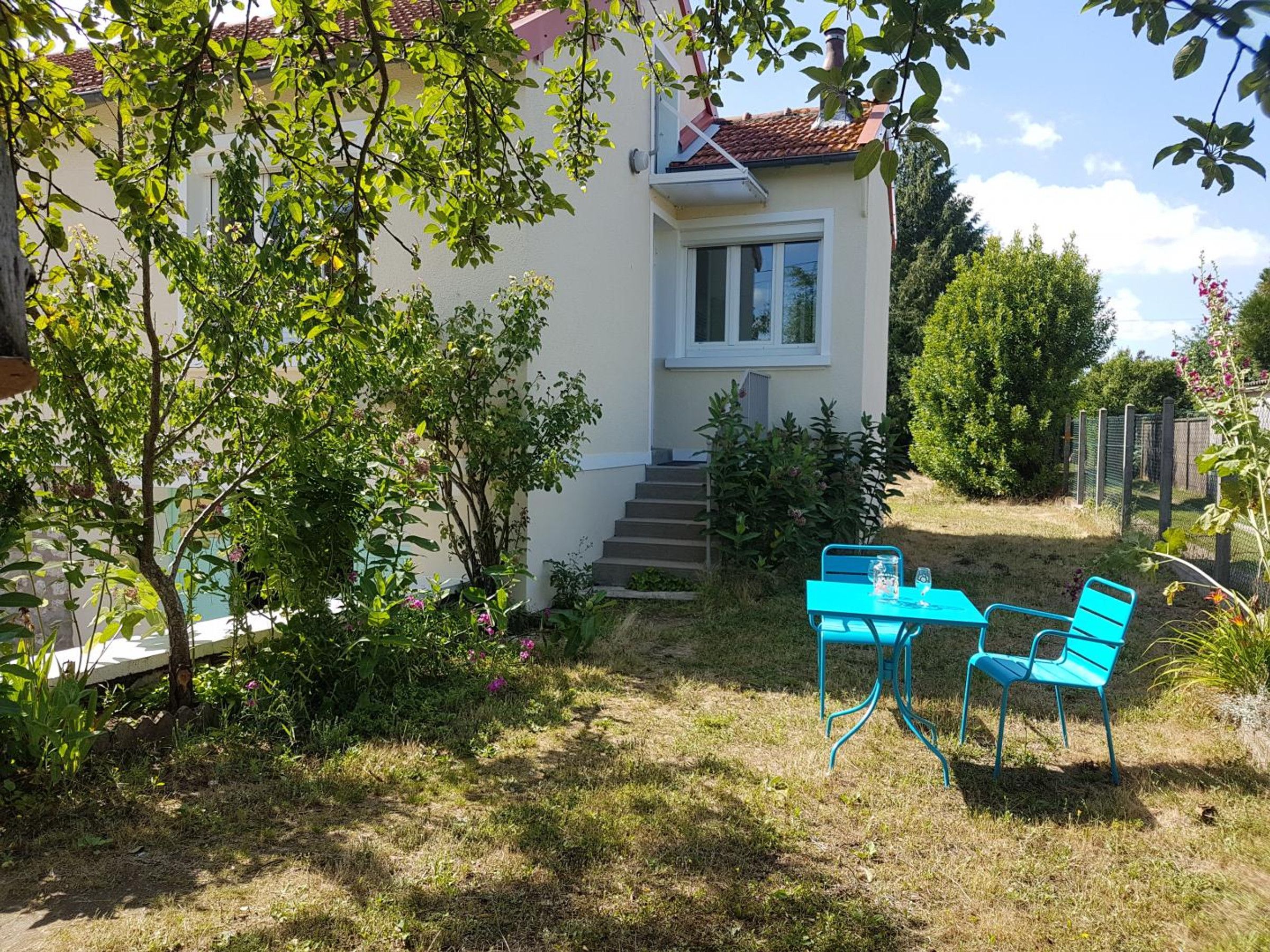 A house with a white exterior, a red-tiled roof, and a small front porch. There are two blue chairs and a table outside.