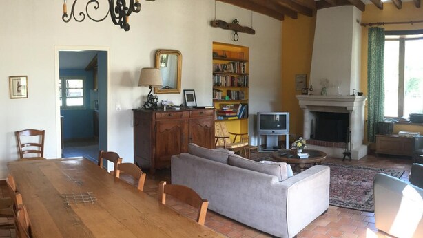 A dining area with a wooden table and chairs, a grey sofa, a fireplace, and a bookshelf with books and a television. - Single-story house with private garden, pets allowed (Villeherviers)