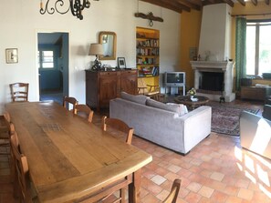 Een eetgedeelte met een houten tafel en stoelen, een grijze bank, een open haard en een boekenkast met boeken en een televisie.