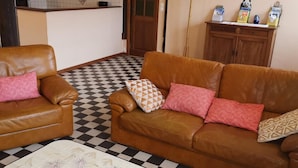Una sala de estar con piso a cuadros, dos sofás de cuero marrón con cojines rosa y estampados, un tapete blanco con motivos florales y un mueble de madera con libros y figuritas.