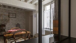 A dining area with a wooden table and chairs, a white brick wall, and a window with curtains. - Charming Cottage in Cheverny with Garden and Garden Furniture (Cheverny)