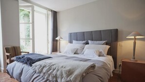 Een net opgemaakt bed met een grijs hoofdbord, twee nachtlampjes en uitzicht op groen.