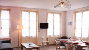 A well-lit living room with a sofa, a dining table set for four, and a TV mounted on the wall.