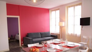 A modern living room with a dining area, a grey sofa, a flat-screen TV, and a large window with curtains.