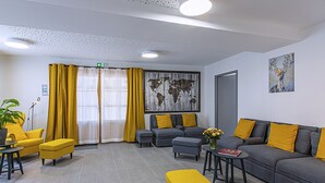 Un salon moderne avec un canapé gris, des coussins jaunes et un fauteuil gris. On y trouve une table basse avec un livre et un vase de fleurs. Un grand tableau est accroché au mur, et une plante en pot est posée sur une table.