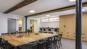 Un espace repas moderne avec une table en bois et des chaises noires, une cuisine en arrière-plan et une enseigne indiquant « Bar-Jeux Restaurant la Croix blanche ».