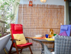 A small outdoor seating area with a bamboo wall, a red and yellow cushioned chair, a wicker chair, and a table with various items.