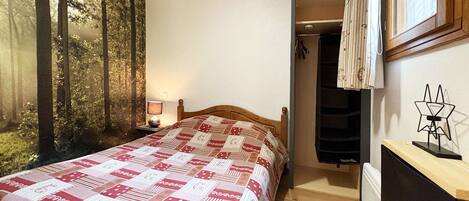 Une chambre à coucher comprenant un lit, une table de chevet, une lampe fixée au mur, une armoire et une fenêtre avec des rideaux.