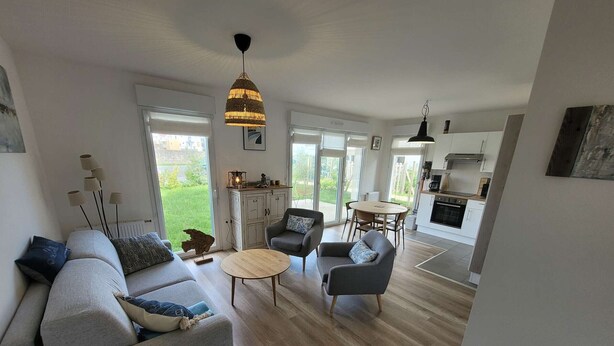 Ein modernes Wohnzimmer mit grauem Sofa, zwei Sesseln, einem Couchtisch und einem Esstisch. Große Fenster bieten einen Blick ins Freie.