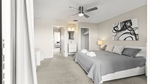 Camera da letto con un letto, una ventola a soffitto, un quadro e un bagno visibile attraverso una porta aperta.