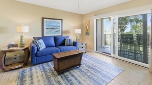 A living room with a blue sofa, a coffee table, and a ceiling fan. There is a large window with a view of palm trees. - Fantastic views from this delightful unit! (St. Augustine)