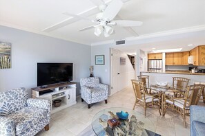 A living room with a television, patterned sofas, a ceiling fan, and a dining area with wicker chairs and a glass table.