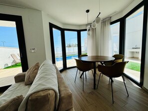 A modern living room with a sofa, a dining table with chairs, and large windows offering a view of a swimming pool and greenery. - Villa in Finestrat Privezwembad, Zee- en Bergzicht (Finestrat)