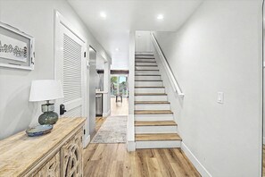 Een hal met een trap naar een balkon, een deur met een raam en een decoratieve schaal op een houten kast.